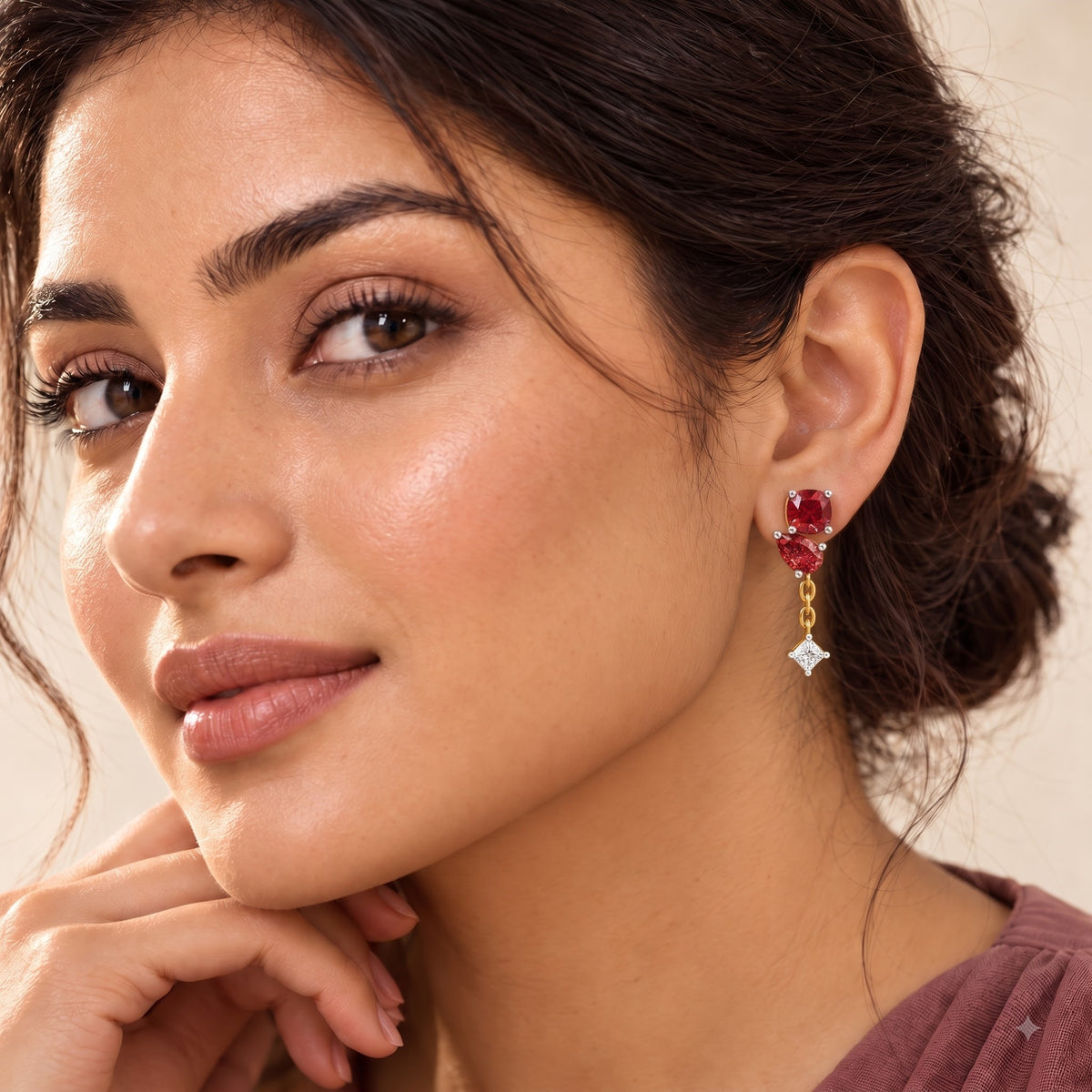 A close-up of a woman wearing a gold drop earring with red gemstones and a dangling clear stone, shown against a soft beige background.