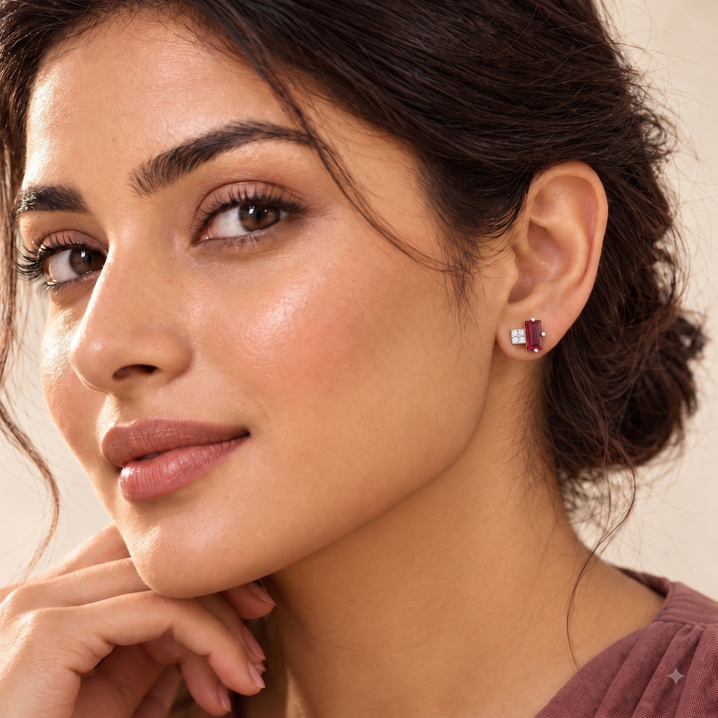 A close-up of a woman wearing a small gold heart-shaped stud earring with a clear central stone. The image is tightly cropped on her face, ear, and hand against a soft beige background.