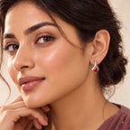 A close-up of a woman wearing a small gold heart-shaped stud earring with a clear central stone. The image is tightly cropped on her face, ear, and hand against a soft beige background.