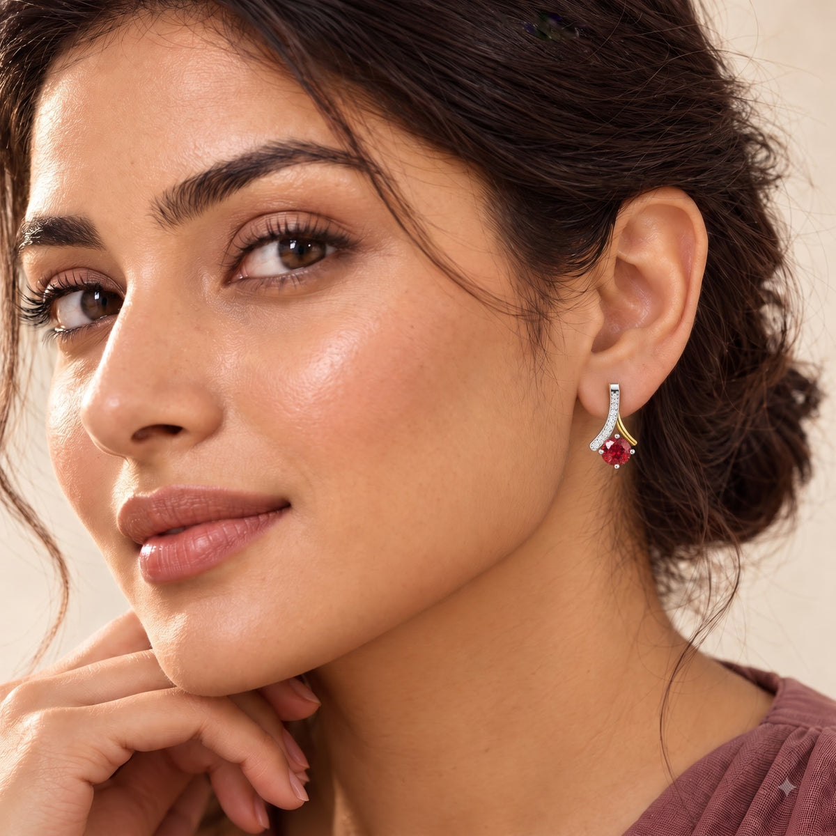 A close-up of a woman wearing a small gold heart-shaped stud earring with a clear central stone. The image is tightly cropped on her face, ear, and hand against a soft beige background.